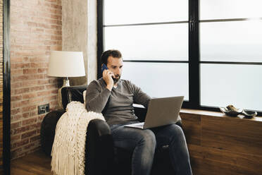 Mature man sitting by window, using laptop, sitting in armchair and talking on the phone - DGOF01088