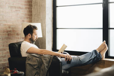 Mature man sitting barefoot in armchair, reading bok - DGOF01094