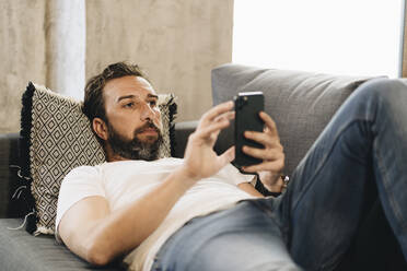 Älterer Mann liegt auf der Couch und benutzt sein Smartphone - DGOF01096