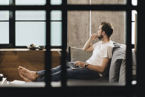 Matur man sitting on couch with laptop on lap, looking away, thinking - DGOF01103