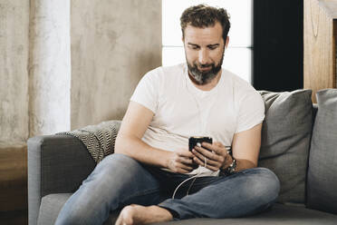 Mature man sitting on couch, using smartphone - DGOF01104
