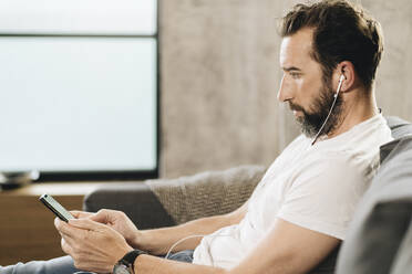 Mature man sitting on couch, using smartphone and earphones - DGOF01108