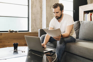 Mature man sitting on couch, using laptop, holding papers - DGOF01112