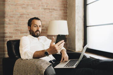 Mature man sitting in armchair, using laptop, holding smartphone - DGOF01117