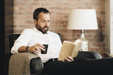 Mature man sitting in armchair, reading book, drinking coffee - DGOF01120