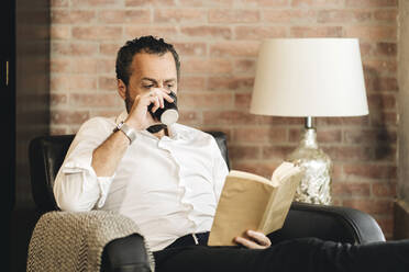 Mature man sitting in armchair, reading book, drinking coffee - DGOF01121