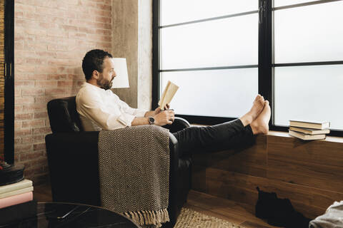 Älterer Mann sitzt im Sessel am Fenster und liest ein Buch, lizenzfreies Stockfoto