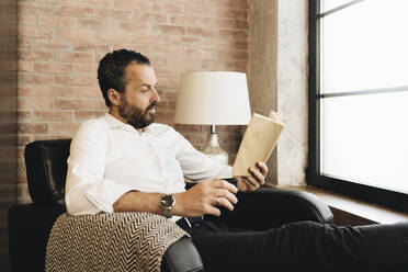 Mature man sitting in armchair by window, reading book - DGOF01123