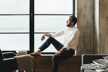 Mature man sitting barefoot on window sill, using smartphone and earphones - DGOF01127