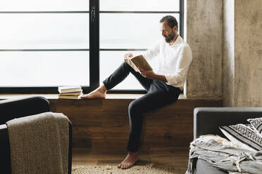 Mature man sitting barefoot on window sill, reading book - DGOF01130