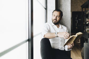 Mature man sitting on window sill, reading book - DGOF01132