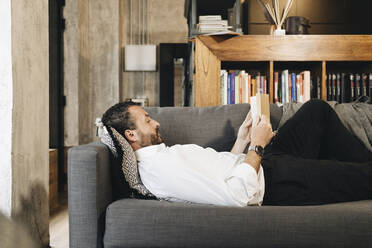 Mature man lying on couch, relaxing, reading book - DGOF01133