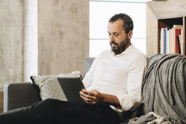 Mature man sitting on couch, using digital tablet - DGOF01138
