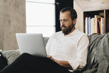 Älterer Mann sitzt auf der Couch und benutzt einen Laptop - DGOF01140