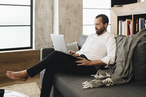 Älterer Mann sitzt auf der Couch und benutzt einen Laptop, lizenzfreies Stockfoto