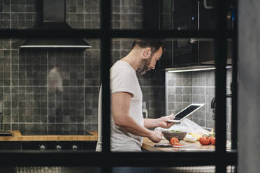 Mature man standing in kitchen, preparing food, using online recipe - DGOF01148