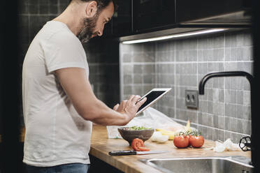 Mature man standing in kitchen, preparing food, using online recipe - DGOF01149