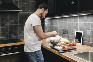 Mature man standing in kitchen, preparing food, using online recipe - DGOF01150