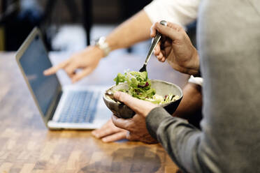Man holding bowl with salad while other one pointing at laptop - DGOF01157
