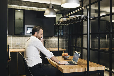 Älterer Geschäftsmann, der in der Küche arbeitet, einen Laptop benutzt und telefoniert, lizenzfreies Stockfoto