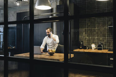 Mature businessman working in kitchen, using laptop, talking on the phone - DGOF01169