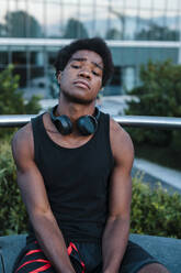 Portrait of a confident young man with headphones sitting in the city - JMPF00038