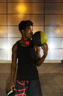 Porträt eines jungen Mannes, der einen Basketball bei Nacht mit Lichtern der Stadt hält - JMPF00051