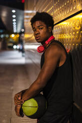 Portrait of a young man holding basketball at night in the city - JMPF00053