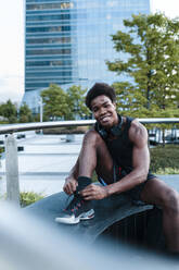 Portrait of a smiling young man with headphones tying his shoes in the city - JMPF00065