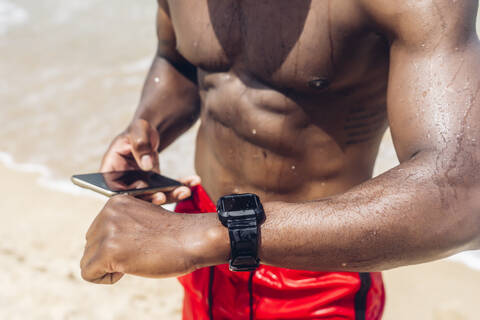 Athletic man with smartphone and smartwatch at beach stock photo