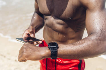 Sportlicher Mann mit Smartphone und Smartwatch am Strand - OCMF01413