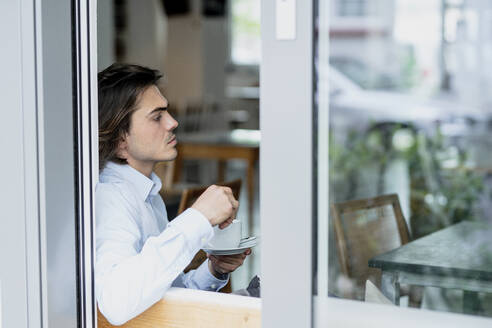 Nachdenklicher Geschäftsmann, der eine Kaffeetasse hält, während er in einem Café sitzt, gesehen durch ein Fenster - KNSF08111