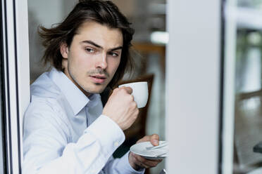 Businessman holding coffee cup looking through window while sitting in cafe - KNSF08113