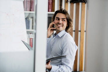 Lächelnder Geschäftsmann, der über sein Smartphone spricht und wegschaut, während er im Büro sitzt - KNSF08138