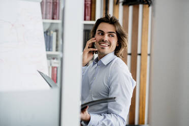 Smiling businessman talking over smart phone looking away while sitting in office - KNSF08138