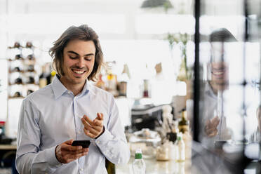 Lächelnder Geschäftsmann, der ein Smartphone benutzt, während er in einem Café steht - KNSF08141