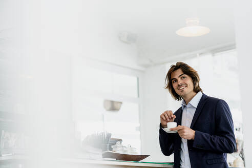 Lächelnder männlicher Unternehmer schaut weg, während er eine Kaffeetasse in einem Café hält - KNSF08148