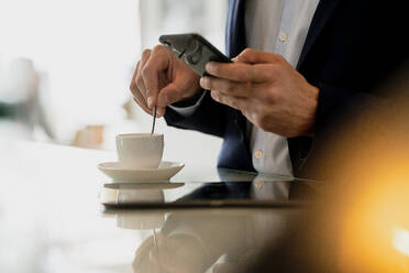 Nahaufnahme eines männlichen Berufstätigen, der sein Smartphone benutzt, während er Kaffee auf dem Tresen eines Cafés mischt - KNSF08152