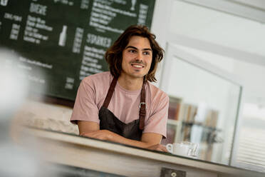 Lächelnder männlicher Besitzer, der wegschaut, während er am Tresen eines Cafés steht - KNSF08157