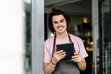 Lächelnder männlicher Besitzer, der ein digitales Tablet benutzt, während er am Eingang eines Cafés steht - KNSF08160