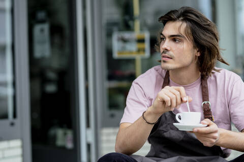 Nachdenklicher männlicher Besitzer hält Kaffeetasse, während er vor einem Café sitzt, lizenzfreies Stockfoto