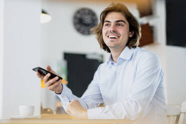 Cheerful businessman holding smart phone while sitting in cafe - KNSF08182