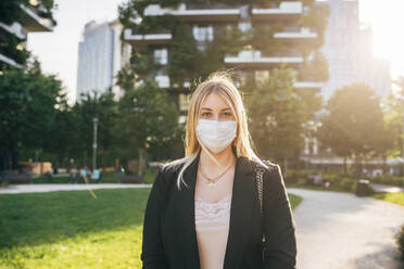 Businesswoman wearing mask standing on footpath in city during sunny day - MEUF01086