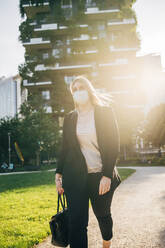 Female business professional wearing mask walking against building in city - MEUF01089