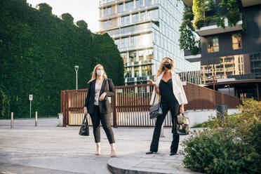 Female business professionals wearing masks standing on street in city - MEUF01093