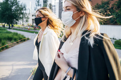 Female colleagues wearing masks walking on footpath in city - MEUF01094