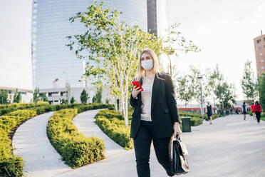 Geschäftsfrau mit Maske, die ein Smartphone hält, während sie auf einem Fußweg in der Stadt spazieren geht - MEUF01095