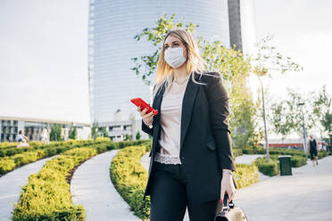 Geschäftsfrau mit Gesichtsmaske, die ein Smartphone hält, während sie auf einem Fußweg in der Stadt spazieren geht - MEUF01099