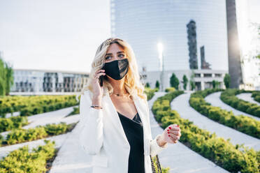 Female entrepreneur wearing face mask talking over smart phone while standing in city - MEUF01104