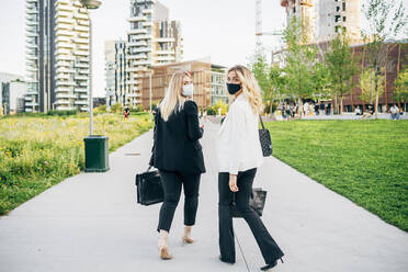 Female coworkers wearing face masks walking on footpath in city - MEUF01106